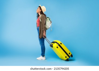 Side View Of Young Black Woman With Backpack And Bright Suitcase Going On Vacation, Traveling Abroad On Blue Studio Background, Copy Space. Tourism, Summer Holidays Concept