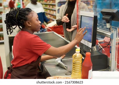 Side View Of Young Black Woman In Uniform Touching Display With Prices Of Food Products While Serving Buyer By Cashier Counter