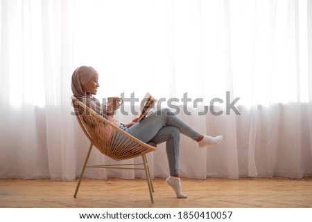 Side view of young black woman in hijab drinking coffee and reading in cozy armchair indoors, copy space. Millennial Islamic woman in traditional headscarf enjoying hot beverage and book at home