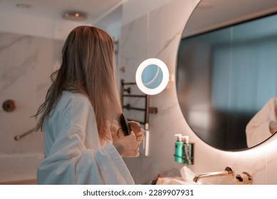 Side view of young blond woman dressed in bathrobe looking in mirror and brushing her hair while standing in modern bathroom at luxurious hotel, luxury vacation concept - Powered by Shutterstock