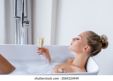 side view of young blonde woman taking bath and holding glass of champagne