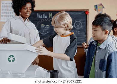 Side view of young boy putting recyclable in waste sorting bin during environmental awareness lesson in multiethnic class - Powered by Shutterstock