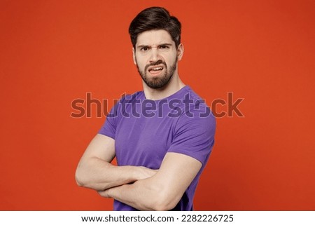 Side view young brunet caucasian man 30s in casual basic purple t-shirt look camera hold hands crossed folded look camera isolated on plain orange background studio portrait. People lifestyle concept