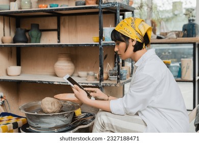 Side view of young brunette asian female artist in headscarf and workwear using digital tablet with blank screen while sitting in ceramic studio at background, craftsmanship in pottery making - Powered by Shutterstock