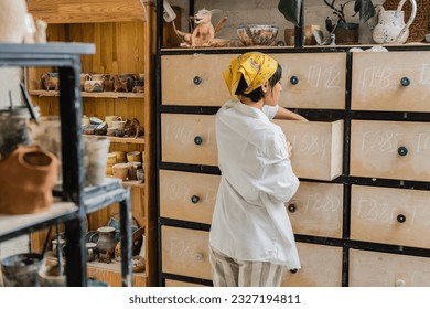 Side view of young brunette asian craftswoman in headscarf and workwear opening cupboard while standing and working in ceramic studio, pottery workshop with skilled artisan - Powered by Shutterstock