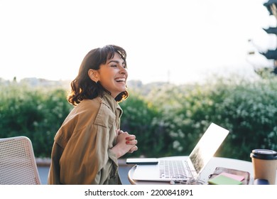 Side view of young brunette smiling happily while sitting at table with laptop working remotely on new web design project in sunny garden