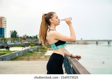 Side View Of Young Brunette Woman Wearing Sports Bra Standing On City Park, Outdoors Drinking Water To Refreshment With Smile On Face. Outdoor Sports, Healthy Life Concepts.