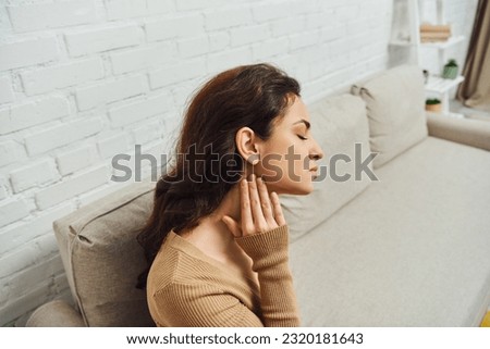 Side view of young brunette woman in brown jumper massaging neck during massage for lymphatic circulation and sitting on couch at home, self-care ritual and holistic wellness practices concept