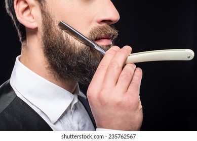 Side View Of A Young Brutal Bearded Man With Straight Razor. Black Background.