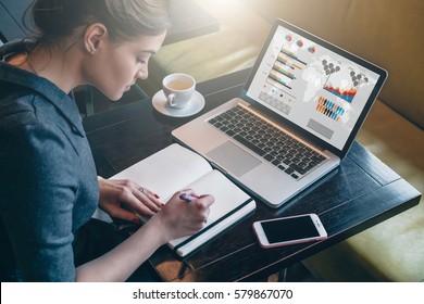 Side view. Young business woman sitting at table and taking notes in notebook.On table is laptop, smartphone and cup of coffee.On computer screen graphics and charts. Student learning online. Blogger. - Shutterstock ID 579867070