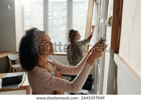 Side view of young businesswoman with paintroller and her colleague painting walls of cafe while renovating interior before opening