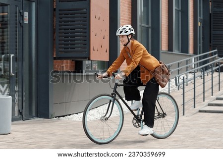 Side view of young businesswoman in quiet luxury attire and protective helmet riding bicycle along modern building of office center after work