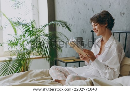 Side view young calm happy woman wear white shirt pajama she lying in bed read book study rest relax spend time in bedroom lounge home in own room hotel wake up dream be lost in reverie good mood day.