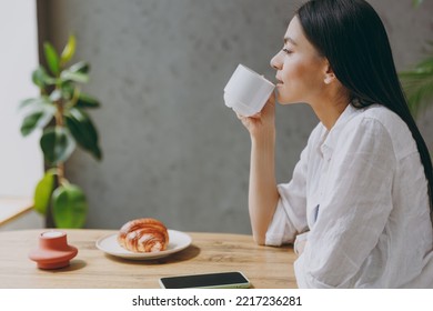 Side view young calm minded latin woman 30s wears white shirt hold cup drink coffee eat croissant sit alone at table in coffee shop cafe restaurant indoors. Freelance mobile office business concept