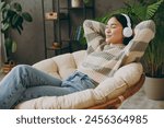 Side view young calm woman wear casual clothes listen music in headphones close eyes sit on armchair stay at home hotel flat rest relax spend free spare time in grey living room indoor. Lounge concept