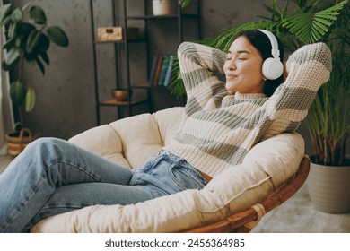 Side view young calm woman wear casual clothes listen music in headphones close eyes sit on armchair stay at home hotel flat rest relax spend free spare time in grey living room indoor. Lounge concept