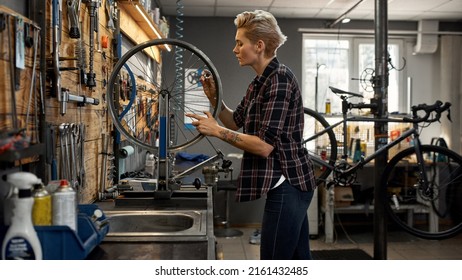 Side view of young caucasian blonde female cycling mechanic checking bicycle wheel spoke with bike spoke key in workshop. Bike service, repair and upgrade. Garage interior with tools and equipment