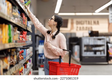 Side view of young Caucasian female student holds basket and reach hand to takes out grocerie from top shelf. Shopping in supermarket and grocery store. - Powered by Shutterstock