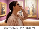 Side view of young Caucasian woman wearing glasses and looking at exhibition. Masterpieces in background. Concept of Museum Day and cultural education.