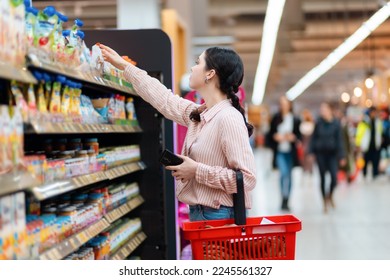 Side view of young Caucasian woman reaches hand to top shelf, holds cellphone and cart. Shelves with food in background. Concept of shopping in grocery store. - Powered by Shutterstock