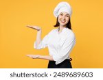 Side view young chef cook baker woman wear white shirt black apron uniform toque chefs hat gesturing demonstrating size with workspace isolated on plain yellow background studio. Cooking food concept