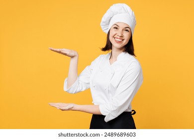 Side view young chef cook baker woman wear white shirt black apron uniform toque chefs hat gesturing demonstrating size with workspace isolated on plain yellow background studio. Cooking food concept
