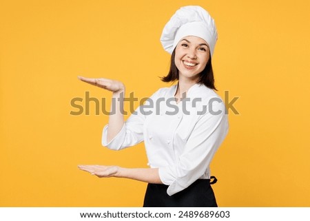 Side view young chef cook baker woman wear white shirt black apron uniform toque chefs hat gesturing demonstrating size with workspace isolated on plain yellow background studio. Cooking food concept