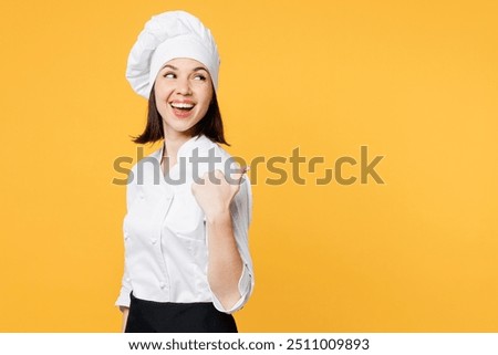 Side view young chef cook baker woman she wear white shirt black apron uniform toque chefs hat point thumb finger aside on area mock up isolated on plain yellow background studio. Cooking food concept