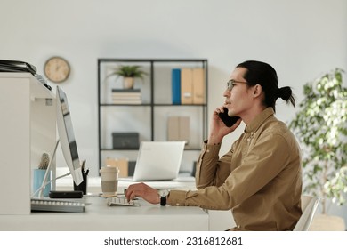 Side view of young confident chief executive officer or director of company talking to business partner on smartphone while sitting by workplace