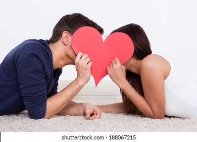Side view of young couple hiding behind heart shape while lying on rug at home