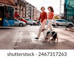 Side view of young couple, man and woman, are walking through city through pedestrian crossing with their dog. Sunset light in background.