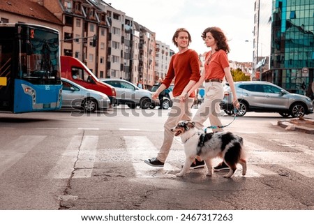 Side view of young couple, man and woman, are walking through city through pedestrian crossing with their dog. Sunset light in background.