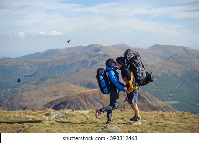 Side view of young couple on backpacking trip standing on mountain summit, kissing and enjoying the view of beautiful landscape against mountain background. Hiking gear/equipment.
