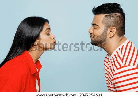 Side view young couple two friend family Indian man woman wear red casual clothes t-shirts together kissing each other with closed eyes while stand face to face isolated on plain blue color background