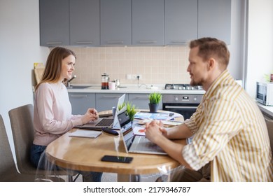Side view of young couple working from home on laptops sitting together at table. Wife and husband discussing business while working remotely or as freelancers