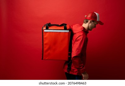 Side view of young courier in a red uniform and with a thermo bag on color red background, space for text. Food delivery service.