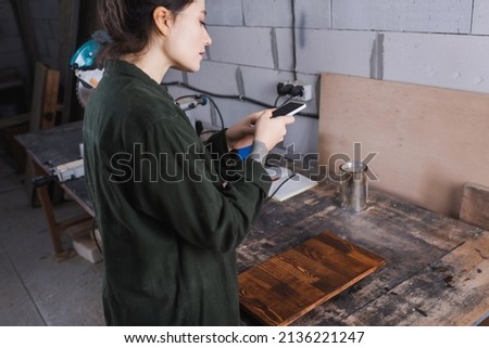 Side view of young craftswoman using smartphone near wooden plank in workshop