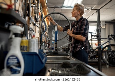 Side view of young european blonde female cycling technician checking bicycle wheel spoke with bike spoke wrench in workshop. Bike service, repair and upgrade. Garage interior with tools and equipment