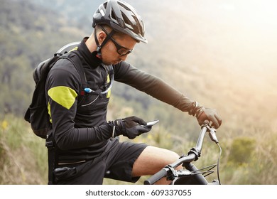 Side view of young European rider standing on top of hill, holding mobile phone, using online application for searching GPS coordinates while riding mootor-powered electric bike in forest on sunny day - Powered by Shutterstock