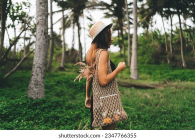 Side view of young faceless female with handbag and straw hat standing in green park while enjoying lush green grass and tress against blurred forest during holiday