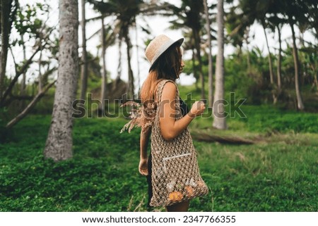 Side view of young faceless female with handbag and straw hat standing in green park while enjoying lush green grass and tress against blurred forest during holiday