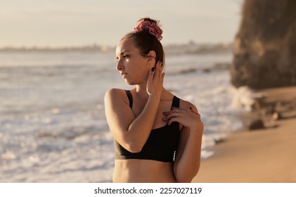 Side view of young female athlete in sportswear standing on sandy beach near waving sea and looking away - Shutterstock ID 2257027119