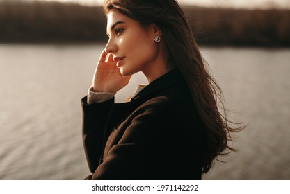 Side view of young female in black coat looking away thoughtfully while standing near river and enjoying sunset in autumn countryside - Powered by Shutterstock