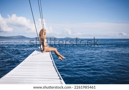 Side view of young female blondie in bikini relaxing on white wooden pier and looking away while sitting with barefoot near sea water and enjoying sunlight of summer