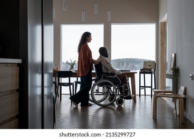 Side View Of Young Female Caregiver Pushing Wheelchair With African American Man While Moving Along Spacious Living Room