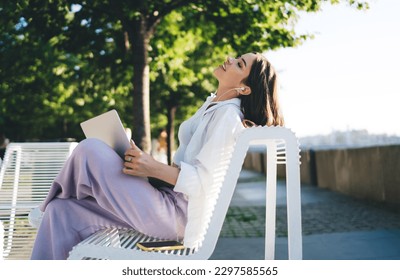 Side view of young female in casual clothes sitting on bench and resting in park on sunny day while looking away dreamily during work break - Shutterstock ID 2297585565