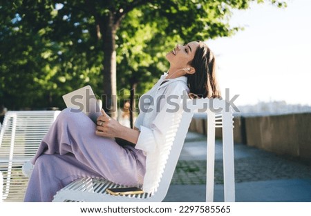 Side view of young female in casual clothes sitting on bench and resting in park on sunny day while looking away dreamily during work break