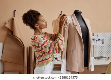 Side view of young female fashion designer sewing handmade decoration on beige blazer hanging on dummy while standing in workshop