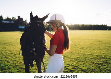 Side View Of Young Female Jockey Wearing Helmet And Red Polo T Shirt Standing Near Chestnut Horse Against Cloudless Sky On Green Field During Training