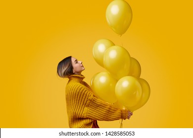 Side view of young female in knitted sweater looking up and releasing bunch of bright balloons during birthday party against yellow background